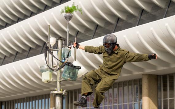 A performer with the astronaut balances precariously on a metal frame with plants