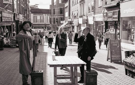 One man drinking while sat at a wooden table in the middle of a street as another man talks to a crowd