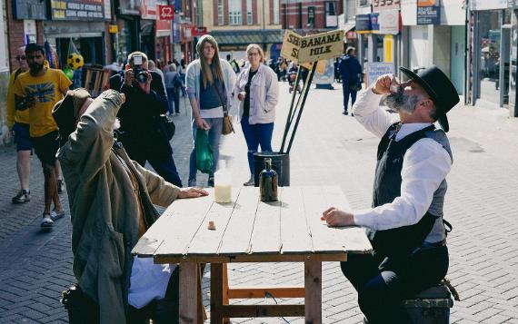 Two men drinking while sat at a wooden table in the middle of a street