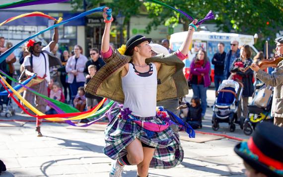 A lady in a purple plaid skirt and tweed jacket dances with colourful ribbons