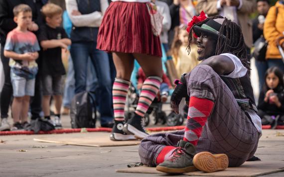 A smiling man in tweed trousers and a waistcoat sits on the floor during a dance routine