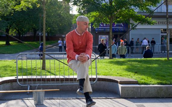 A man in a red shirt sits perfectly balanced on a tight rope in front of trees and grass