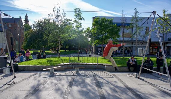 A man in a red shirt moving around on a tight rope in front of an audience next to greenery