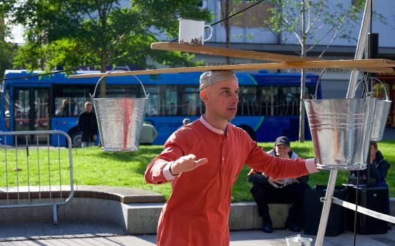 A man in a red shirt balances multiple wooden boards with buckets on the end on his head