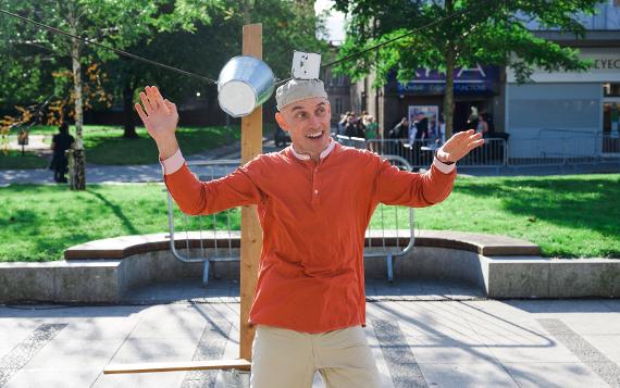 A man in a red shirt smiling to the audience balances a metal cup on his head in front of greenery