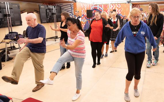 A group of young and old people kick their feet up while swing dancing on a dance floor