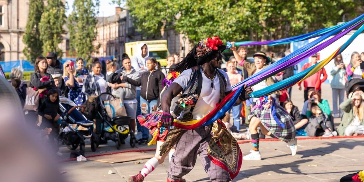 Encounter Festival Folk Dance Remix A man with a flowery hat and baggy clothes dances around a pole holding a brightly coloured long sheet