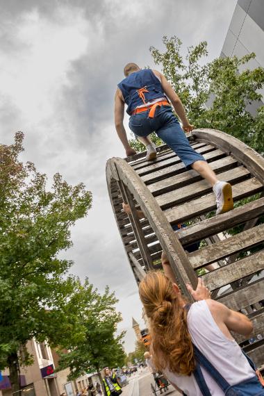 A performer climbs on top of a giant wooden wheel in an art performance