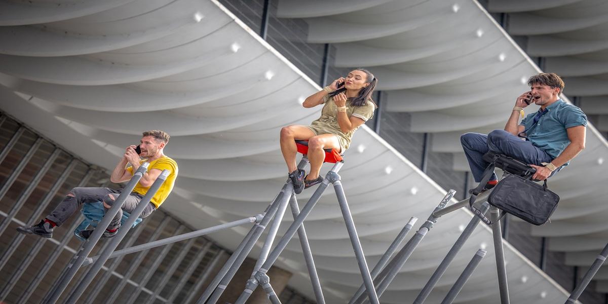 Three people sat on scaffolding on their phones during the performance WILD at Encounter 2024.