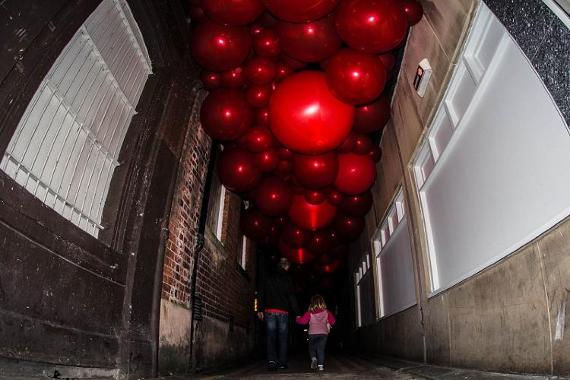 Man holding daughter's hand as they walk down dark alleyway lit by large, round, red balloons above them.